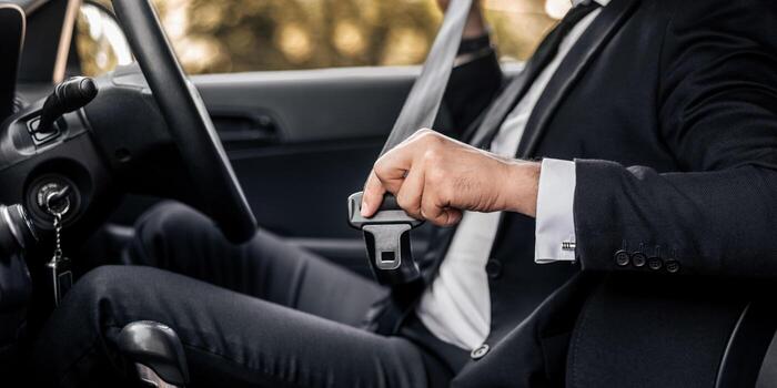 A person in a dark business suit seated in a car, fastening the seat belt buckle before setting off.