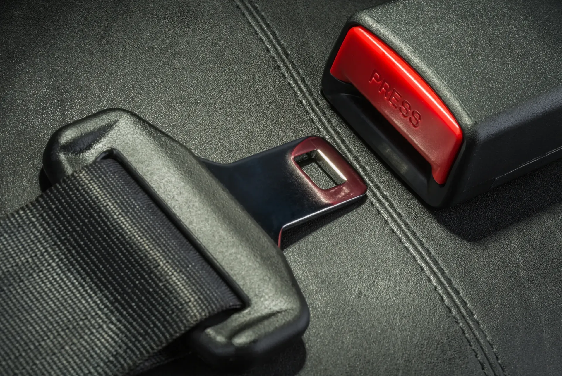 Close-up of an automotive seat belt tongue plate resting beside a black buckle receiver with a red press-release button on a stitched leather seat.