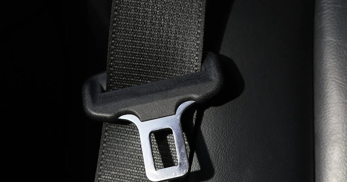 Close-up of black woven seat belt webbing threaded through a moulded guide with the polished metal tongue plate below it.